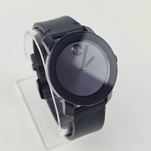 May include: A dark blue wristwatch with a black leather band. The watch face is a gradient of dark blue and gray with silver hands and a single dot at the 12 o'clock position. The band is stitched with blue thread.