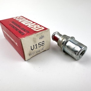 May include: A silver-colored PCV valve next to a red and white cardboard box. The box has the word "STANDARD" in red letters and the text "PROFESSIONAL QUALITY AUTOMOTIVE PARTS". The box also has the text "U158 PCV VALUE".