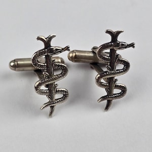 May include: A pair of silver-toned metal cufflinks featuring a detailed design of a snake coiled around a staff. Each cufflink has a polished finish with textured scales on the snake and a standard hinged toggle closure on the back for secure fastening.