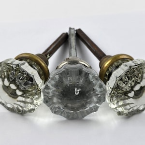 May include: Three clear glass door knobs with faceted surfaces and brass-colored rings. The knobs have a vintage aesthetic, with aged metal stems. The knobs are arranged on a white background, showcasing their intricate design and potential for home decor.