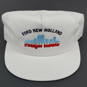 May include: White baseball cap with an embroidered logo that says "Ford New Holland Parts Mart" with a blue and red cityscape graphic.