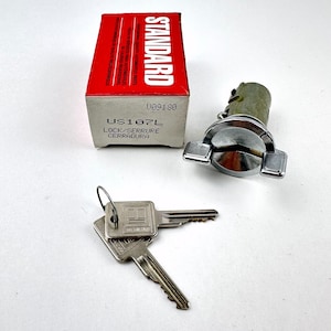 May include: A car ignition lock set with two keys and a box. The box is red and white with the word "STANDARD" printed on it. The keys and lock are silver in color.
