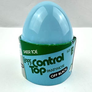 May include: A blue plastic egg-shaped container with a green and white label that reads "Leggs Control Top Pantyhose Off Black Sheer Toe Size B".