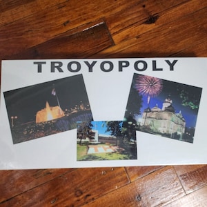 Puede incluir: Una caja blanca con el texto "TROYOPOLY" en letras negras. La caja tiene tres imágenes: una fuente de noche, un edificio con fuegos artificiales y un edificio con un césped verde.