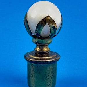 May include: A decorative bottle stopper with a white orb top, framed by ornate, aged metalwork. The base is a cylindrical shape with a green patina, and the overall design has a vintage aesthetic.