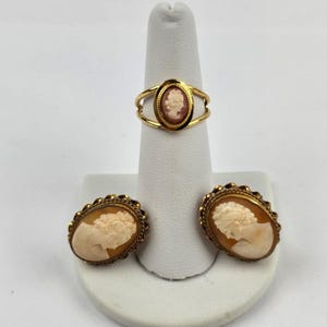 May include: A gold ring featuring a small oval cameo with a profile portrait and a pair of matching gold oval earrings, each displaying a carved cream-colored profile portrait against a warm tan background with a textured gold border.