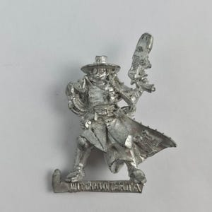 May include: A silver-colored miniature figure of a character in a hat and long coat, holding a futuristic gun. The figure is in a dynamic pose, as if in action. The base of the figure has the text "WITCHHUNTER.R.I.N.A."