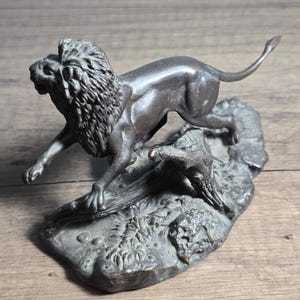 May include: A bronze-colored lion sculpture in a dynamic pose, leaping over a textured base. The lion's mane is detailed, and the sculpture has a weathered appearance, suggesting an antique or vintage piece. The sculpture is on a wooden surface.