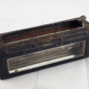 Puede incluir: Una caja rectangular de metal negro con una ventana de vidrio. La caja tiene un interior marrón oxidado y está asegurada con tornillos.