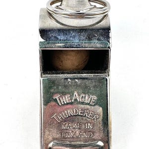 May include: A silver-colored Acme Thunderer whistle. The whistle has a ring for attachment and the text "THE ACME THUNDERER MADE IN ENGLAND" is engraved on the front. The whistle is made of metal and has a small wooden ball inside.