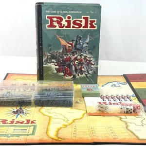 Könnte beinhalten: Ein Risk-Brettspiel-Set mit Spielbrett, Spielfiguren, Würfeln und einer Box. Die Box zeigt den Spieletitel und eine Grafik von Soldaten. Das Spielbrett ist geöffnet und zeigt eine Weltkarte.