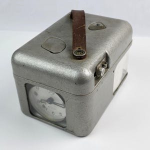 May include: A vintage silver-toned metal clock with a leather handle. The clock face is visible through a glass window. The clock has a rectangular shape with rounded edges. The clock face displays the time with black hands and numerals.