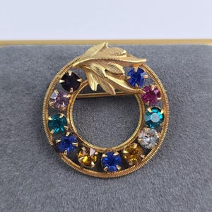 May include: A gold-toned brooch with a circular design, featuring a ring of colorful gemstones. A gold leaf design is at the top. The gemstones are in various colors, including blue, purple, and yellow. The brooch is set against a gray background.