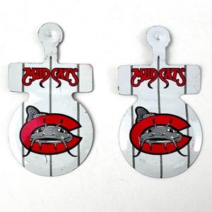 May include: Two white metal Mud Cats logo pins. Each pin features a red letter "C" with a catfish graphic, and the words "Mud Cats" in red. The background has vertical gray stripes.