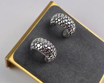 May include: A pair of silver-colored hoop earrings with a textured, bubble-like design. The earrings are displayed on a gray velvet surface set within a gold-colored frame. The earrings have a rounded shape and a polished finish.