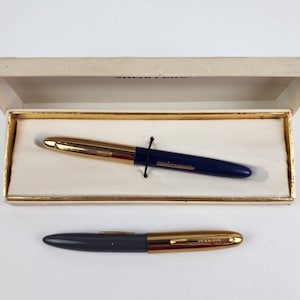 Może przedstawiać: Dwa pióra wieczne w białym pudełku. Górne pióro jest niebieskie z pozłacanymi zdobieniami. Dolne pióro jest szare z pozłacanymi zdobieniami.