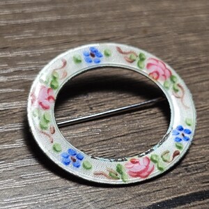 May include: A vintage circular brooch with a floral design. The brooch features a white background with pink roses, blue flowers, and green leaves. The brooch has a silver pin on the back and is set against a wooden surface.