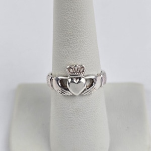 May include: A silver-toned Claddagh ring featuring a central heart shape held by two hands, topped with a small crown. The band is smooth and metallic, showcasing the traditional design elements of a heart, hands, and crown in a polished finish.