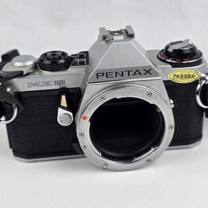 May include: A silver and black Pentax ME Super 35mm film camera body with a lens mount, a film advance lever, a shutter speed dial, and a yellow sticker on the top right side that reads PASSED. The camera includes a black fabric neck strap.