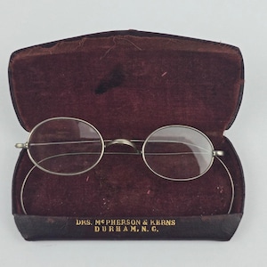 May include: Antique oval-shaped wire-rimmed spectacles with thin metal temples resting inside a dark burgundy velvet-lined case. The case features gold-colored text reading DKS. MCPHERSON & KERNS DURHAM, N. C. printed on the front edge.