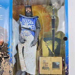 Op de afbeelding: Een ridderactiefiguur in een doorzichtige plastic display case. De figuur draagt een blauw en witte tuniek met een boomontwerp, maliënkolder en een helm. Accessoires: zwaard, schild en een boek met "Scene 24".