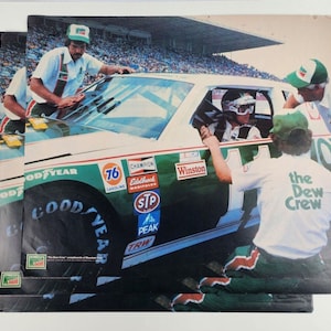May include: A green and white race car with the number 76 on the side. The car is surrounded by three people wearing green and white racing uniforms. The car has several sponsors' logos on the side, including Winston, Champion, Edelbrock, STP, Peak, and TRW. The text "The Dew Crew" is printed on the back of one of the crew member's shirts.