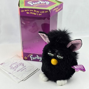 May include: A black electronic Furby toy with white feet, a yellow beak, and pink-lined ears, accompanied by its original purple and green cardboard packaging box featuring the text The more you PLAY with me the MORE I do and an instruction manual.