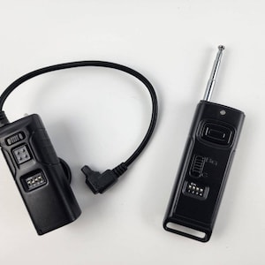 May include: A black wireless flash trigger set with a receiver and a transmitter. The receiver has a small antenna and a dial with multiple settings. The transmitter has a small antenna and a button for triggering the flash.