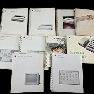 May include: A collection of vintage Apple Macintosh computer manuals, including the Macintosh System Software User's Guide, HyperCard User's Guide, Macintosh 12" RGB Display Owner's Guide, The Applesoft Tutorial, and the Macintosh Guide. The manuals are bound with spiral binding and have white covers with black text.