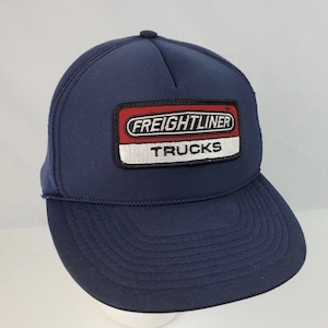 Könnte beinhalten: Eine marineblaue Trucker-Kappe mit einem rot-weiß-schwarzen Patch, auf dem "FREIGHTLINER TRUCKS" steht.