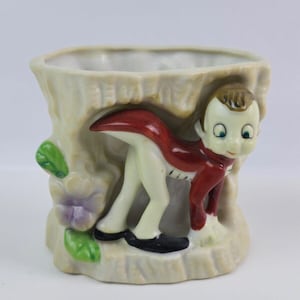 Può includere: Un vaso da fiori in ceramica bianca a forma di ceppo d'albero con una piccola figurina di un ragazzo che spunta da dietro il ceppo. Il ragazzo indossa una giacca rossa, pantaloni bianchi e scarpe nere. Il vaso ha foglie verdi e fiori viola.