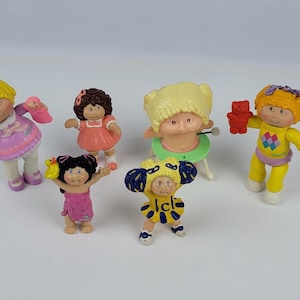 May include: A collection of six Cabbage Patch Kids dolls. The dolls are all wearing different outfits and have different hairstyles. Some of the dolls are holding accessories, such as a baby doll, a teddy bear, and a rubber duck.