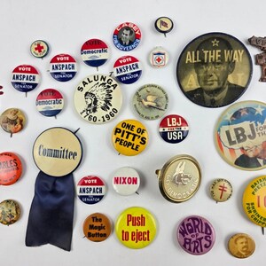 Puede incluir: Una colección de botones y pines de campaña política vintage. Los pines presentan textos como "Vote Anspach", "LBJ for the USA" y "All the Way". Hay varios colores y diseños presentes, incluyendo rojo, blanco, azul y amarillo.