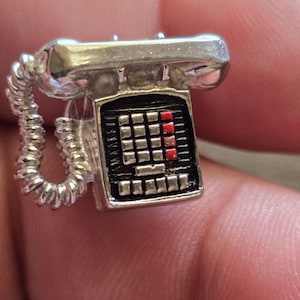 May include: A silver-colored charm in the shape of a vintage telephone. The charm features a detailed design with a coiled cord, a handset, and a keypad with red accents. The charm is small and appears to be made of metal.