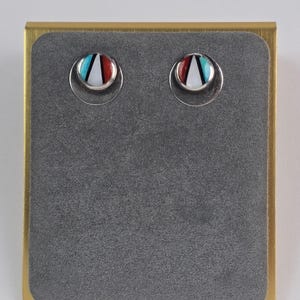 Puede incluir: Un par de pendientes de botón con un diseño circular. Cada pendiente presenta una base blanca con un patrón triangular rojo, turquesa y negro. Los pendientes están engarzados en un respaldo de metal plateado y se exhiben sobre una superficie de terciopelo gris.
