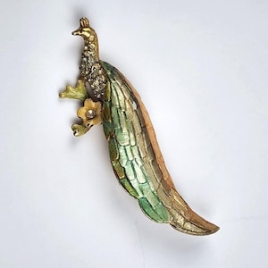 Puede incluir: Broche decorativo de pavo real con cuerpo dorado, plumas de cola verdes y doradas, y cabeza adornada con pedrería. El broche presenta una pequeña flor amarilla y está sobre un fondo blanco.