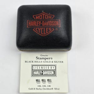 May include: A black rectangular jewelry box with a red Harley-Davidson Motor Cycles logo on the lid, accompanied by a white informational card featuring the text Genuine Stamper Black Hills Gold & Silver Licensed by Harley-Davidson 10K, 12K, 14K.