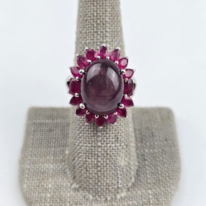 May include: A silver ring featuring a large, oval, cabochon-cut dark pinkish-red gemstone at the center, surrounded by a halo of smaller, pear-shaped, faceted red gemstones set in a prong setting, creating a floral-inspired cluster design.