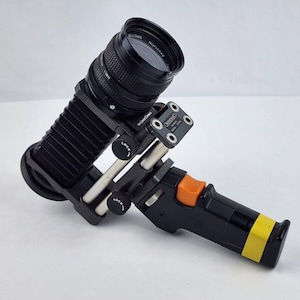 May include: Black bellows camera extension tube with a black lens attached to a black and orange hand grip. The extension tube has a silver lock knob and a silver adjustment knob. The lens has the text "MC ROKKOR 135mm 1:3.5" printed on it.