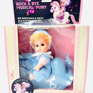May include: A vintage rocking horse toy with a baby doll riding on it. The horse is blue and white and the doll is wearing a blue and white outfit. The box has the words "Rock A Bye Musical Pony" on it.