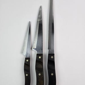 Puede incluir: Tres cuchillos de cocina de acero inoxidable con mangos de madera oscura. Las hojas varían en tamaño y forma, incluyendo un cuchillo de cocina, un cuchillo multiusos y un cuchillo de pan dentado. Cada cuchillo tiene un mango remachado.