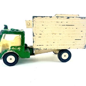 May include: A green toy truck with a white cab and a beige box bed. The truck has a white steering wheel and the words "A.S.E.R.O." printed on the side.