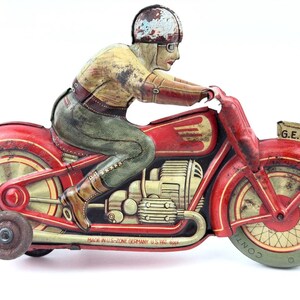 May include: Vintage red and yellow tin toy motorcycle with a rider wearing a helmet and goggles. The motorcycle has a sidecar wheel and the text "G.E. 258" on the front. The text "MADE IN U.S.-ZONE GERMANY U.S. PAT. APP." is visible.