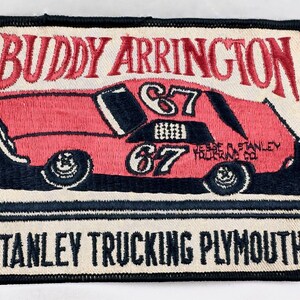 May include: Embroidered patch featuring a red race car with the number 67 on the side. The car is on a black and white background with the text "BUDDY ARRINGTON", "JESSE STANLEY TRUCKING CO.", and "STANLEY TRUCKING PLYMOUTH".