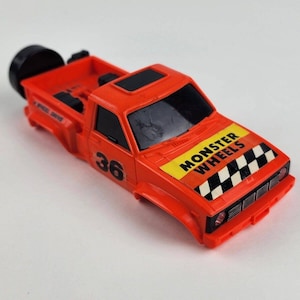 May include: Orange toy truck with black wheels and a black windshield. The truck has a black and white checkered stripe on the hood with the words "Monster Wheels" in yellow letters.
