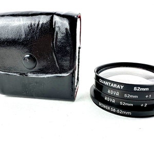 May include: A set of three black camera lens filters in a black leather case. The filters are labelled "QUANTARAY 52mm +1", "HOYA 52mm +1", and "HOYA 52mm +2". The case has a snap closure.
