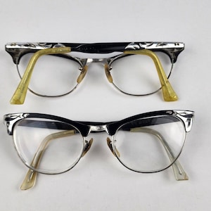 Op de afbeelding: Twee paar vintage cat-eye brillen met zwarte monturen en zilveren accenten. De brillen hebben heldere glazen en gele kunststof pootjes.