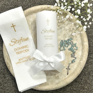 May include: A white pillar candle with a satin ribbon, a white fabric cloth, and a light blue beaded rosary. The candle and cloth are printed with a gold cross and the text Stefan Dominic Tripodi, Baptised 18th May 2025 and 19th May 2024.