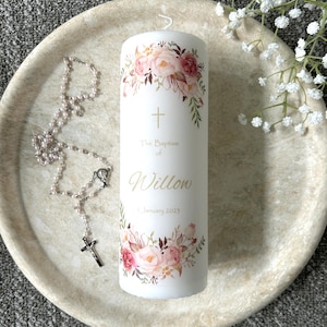 May include: A white cylindrical pillar candle featuring a floral design with pink and peach roses, a gold cross, and the text "The Baptism of Willow 1 January 2023" in gold lettering. A delicate rosary with small pink beads and a silver crucifix is placed beside it.