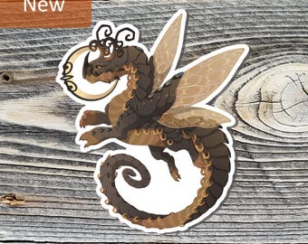 May include: A sticker featuring a brown and tan dragon with wings, a crescent moon, and the word "New" in a brown rectangle. The dragon has a long, curled tail and is set against a wood grain background.